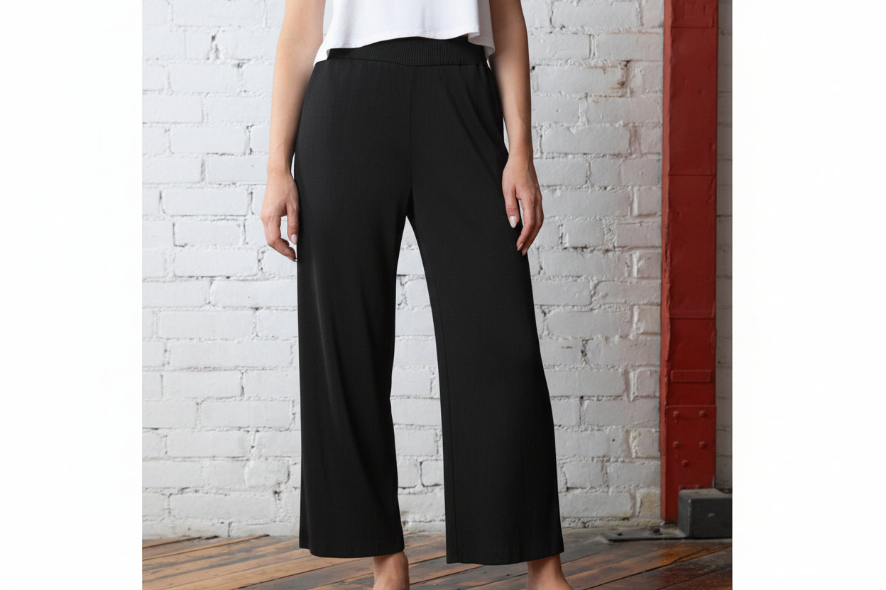 A pair of black mini pleat wide-legged occasion pants with a pull-on design, displayed on a model standing against a brick wall background.