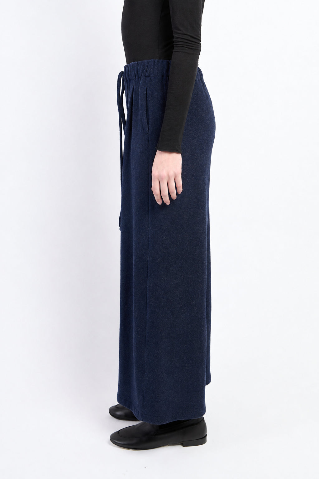 A model shown from the side wearing dark gray wide-leg knit pants with a drawstring waist, a black long-sleeve top, and black shoes against a plain white studio background.
