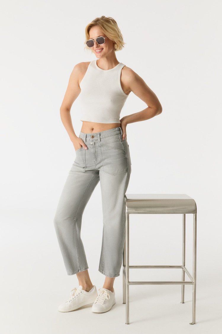 Woman wearing a white sleeveless top and light gray jeans standing next to a stool on a white background