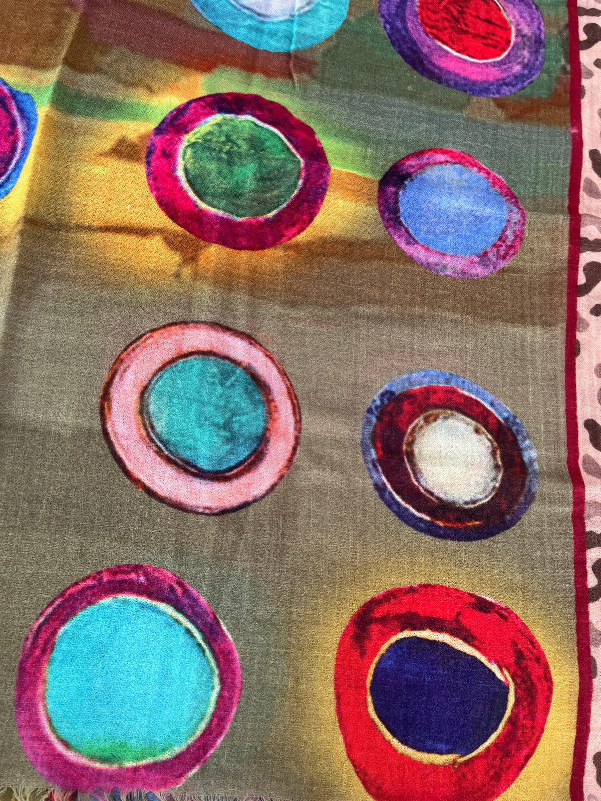 A close-up of a scarf featuring a multicolor abstract pattern of concentric circles in red, pink, blue, green, and purple on a warm brownish-gold background.