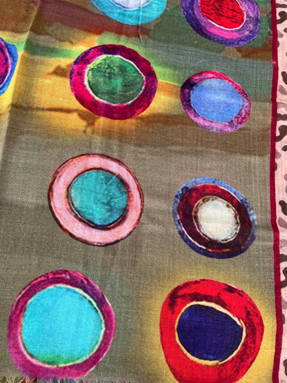 A close-up of a scarf featuring a multicolor abstract pattern of concentric circles in red, pink, blue, green, and purple on a warm brownish-gold background.