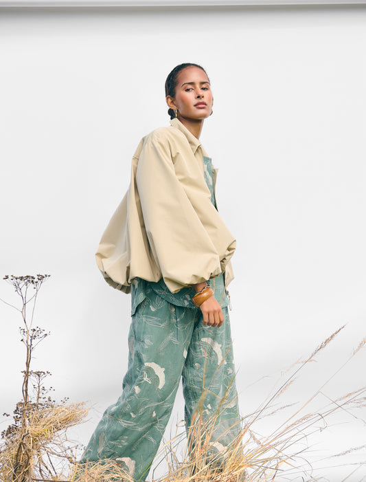 Person wearing a beige jacket and green patterned pants standing in a natural setting with white background