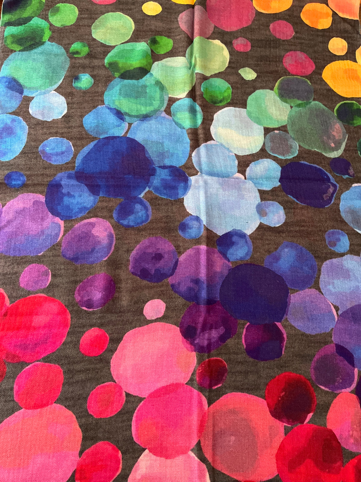 A close-up of a scarf with colorful watercolor-style pattern of overlapping circles in pink, red, purple, blue, green, and yellow on a dark grey background.