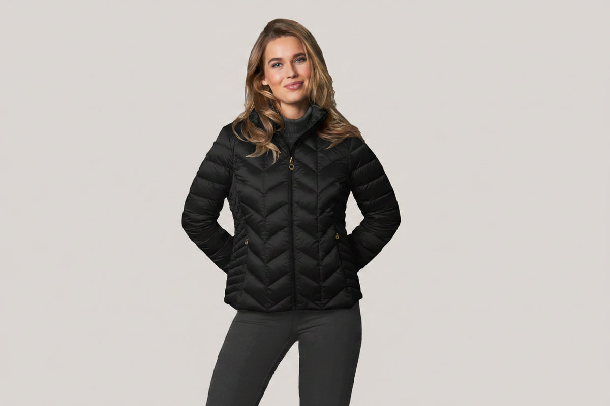 A woman modeling a BLACK chevron-quilted puffer jacket with subtle gold trim on the shoulders and zipper area, paired with dark gray pants, against a solid light gray background.