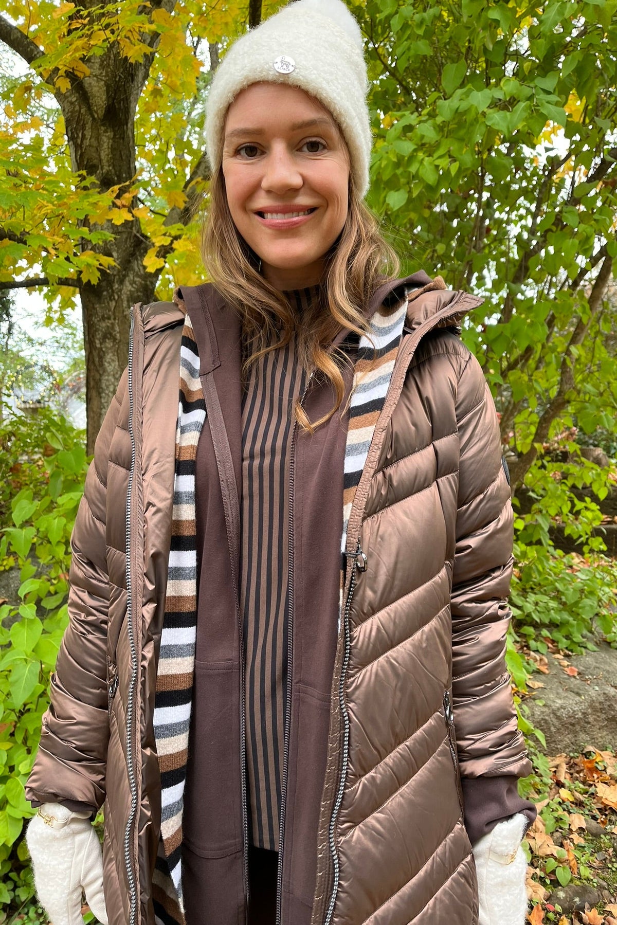 Cari wearing the Junge Phillipa down coat in brown over a Blue Sky Cortes jacket, LUUKAA stripe pullover and HUE leggings, with a Maison Izzy hooded scarf, alpaca pompom hat and LUV & Co. gloves