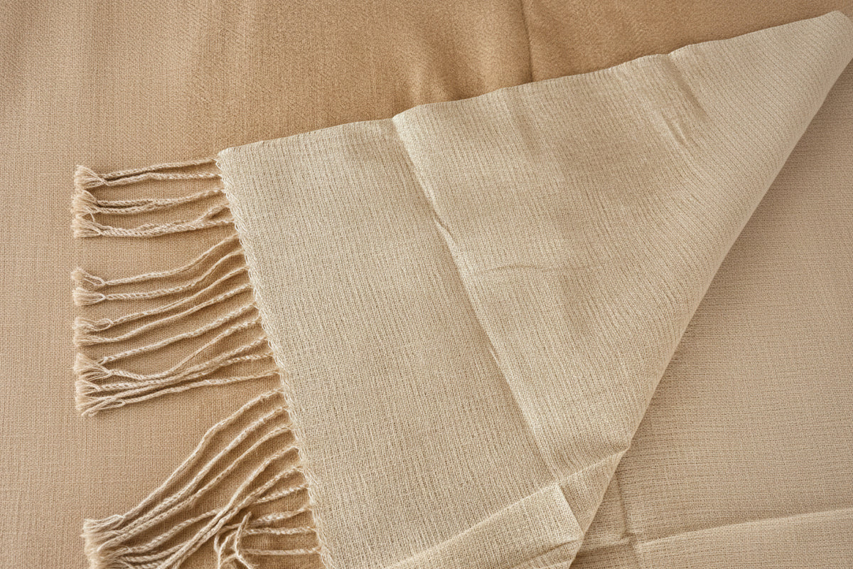 A close-up of a light beige woven throw blanket with fringed edges, draped over a SHOWING REVERSE SIDE of light cream