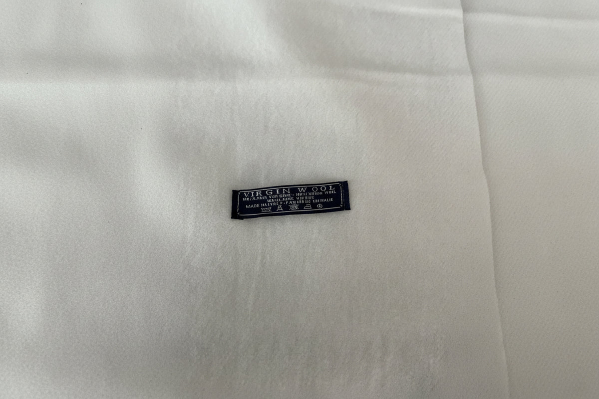 A close-up of white scarf with a small black woven label reading 'VIRGIN WOOL' and additional care text.
