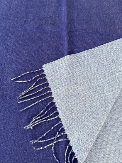 A close-up of a woven scarf  folded to show its texture and fringed edge. The fabric appears in two tones: a rich royal blue on the main body and a lighter icy blue on the folded corner, with twist thread fringe.