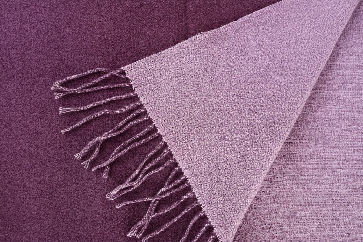 A close-up of a woven scarf  in a  dark purple and reverse side dusty lavender-purple color, showing its textured knit pattern and fringed edges.