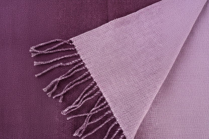 A close-up of a woven scarf  in a  dark purple and reverse side dusty lavender-purple color, showing its textured knit pattern and fringed edges.