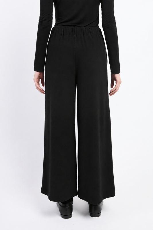 A person shown from the back wearing dark charcoal wide-leg knit pants with an elastic waistband, paired with a black long-sleeve top and black shoes, against a light gray studio background.