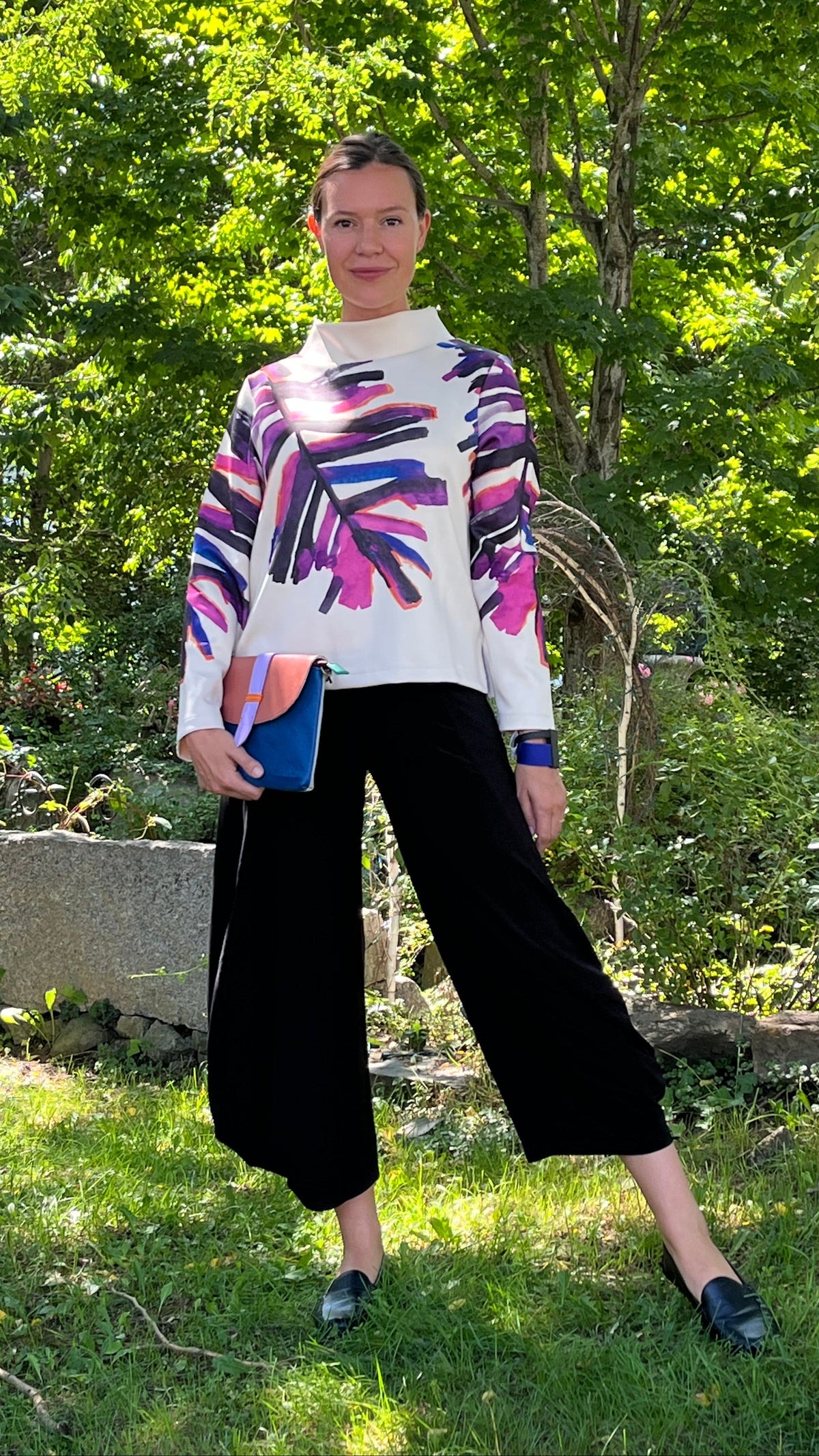 A woman stands outdoors in a grassy garden area with trees in the background, wearing a white turtleneck sweater with a colorful abstract leaf print in purple, pink, and blue, paired with black wide-leg cropped pants and black loafers. She is holding a blue and orange clutch.