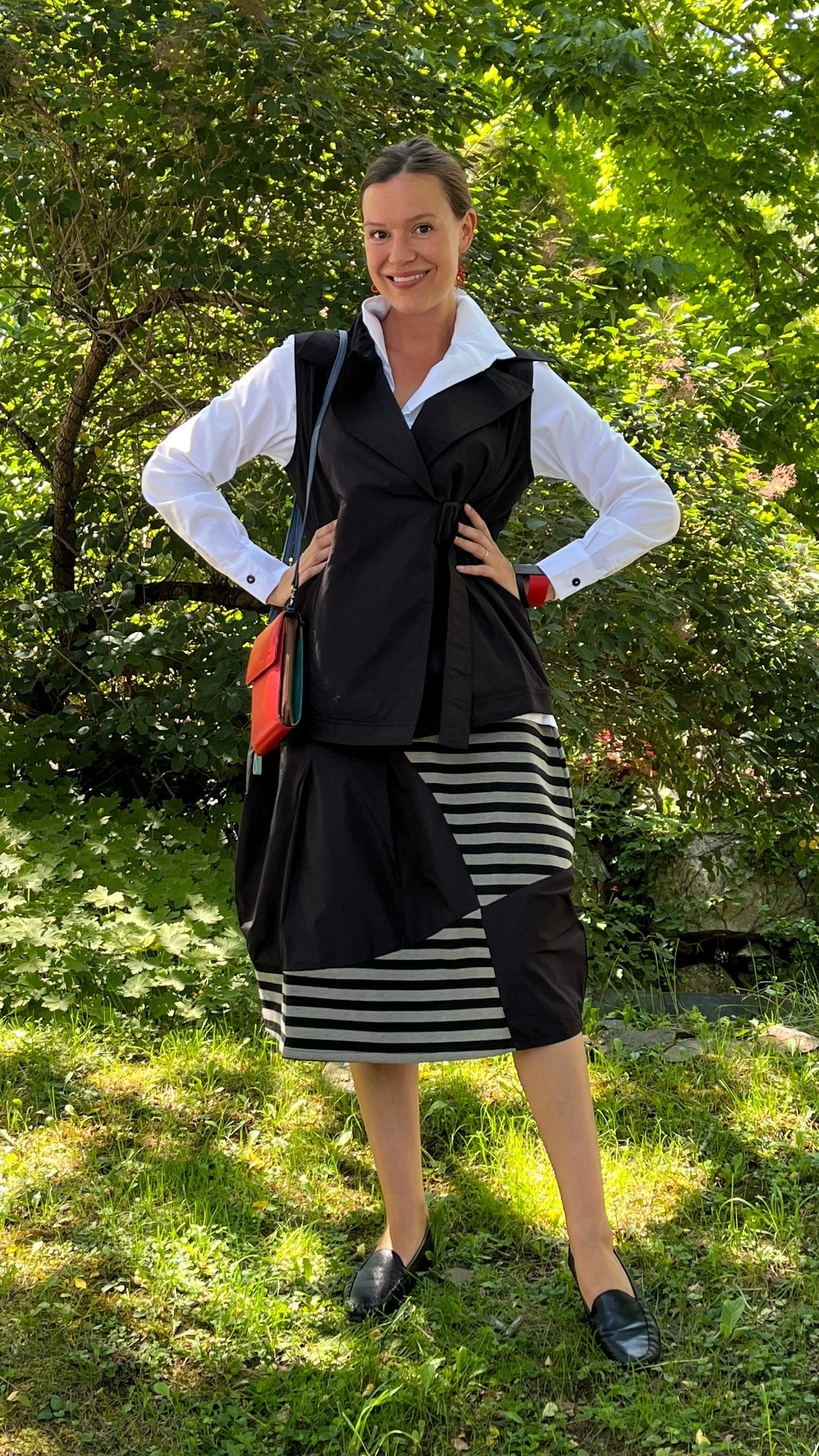 A woman posing outdoors in a garden setting, wearing a black wrap vest over a white collared shirt, a black and white striped skirt, and black loafers, while carrying a red crossbody bag.