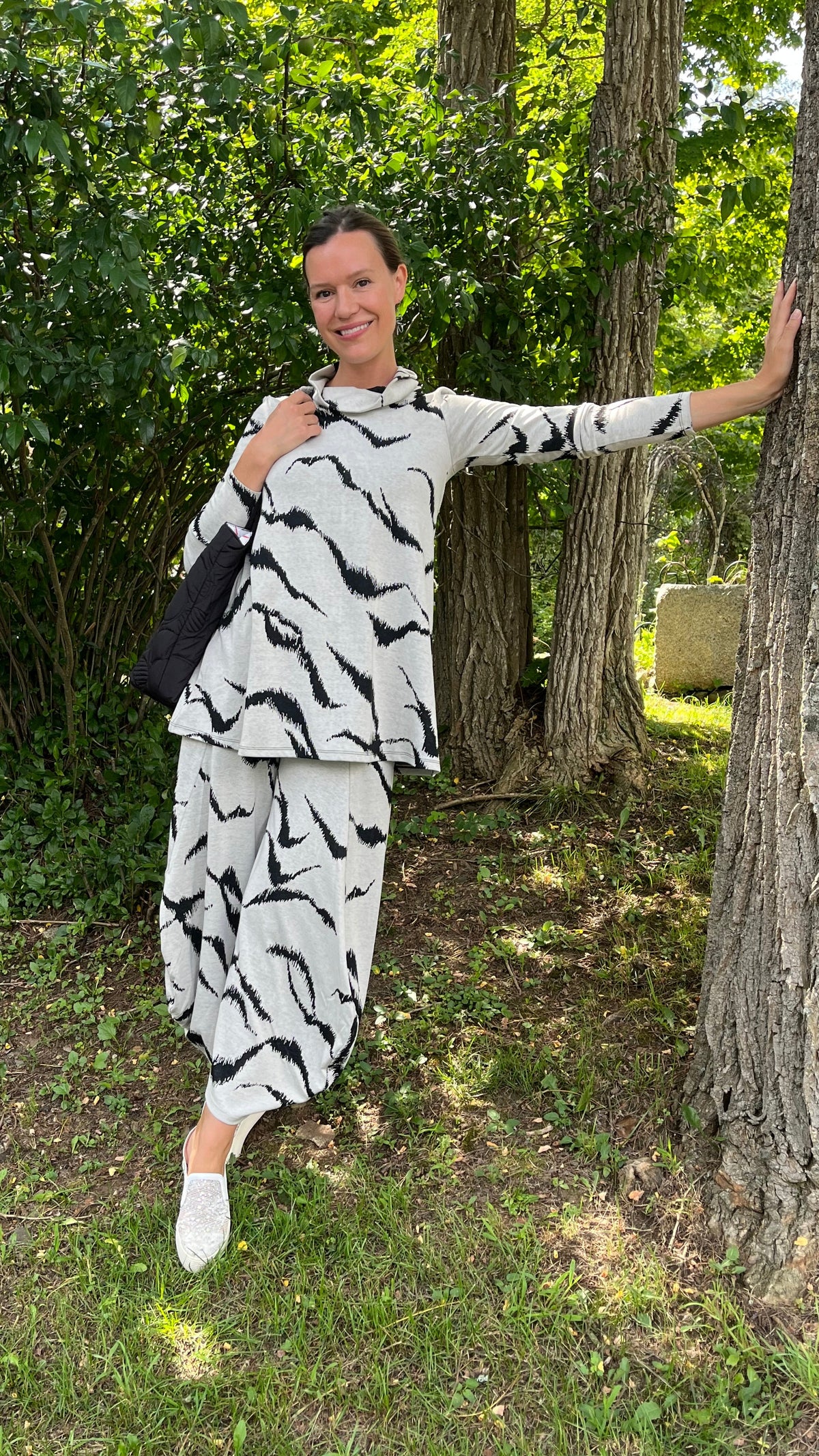 A woman posing outdoors in a wooded area, wearing a matching two-piece set with a black and white zebra-like pattern. The set includes a long-sleeve top and wide-leg pants, paired with white slip-on shoes and a black shoulder bag.
