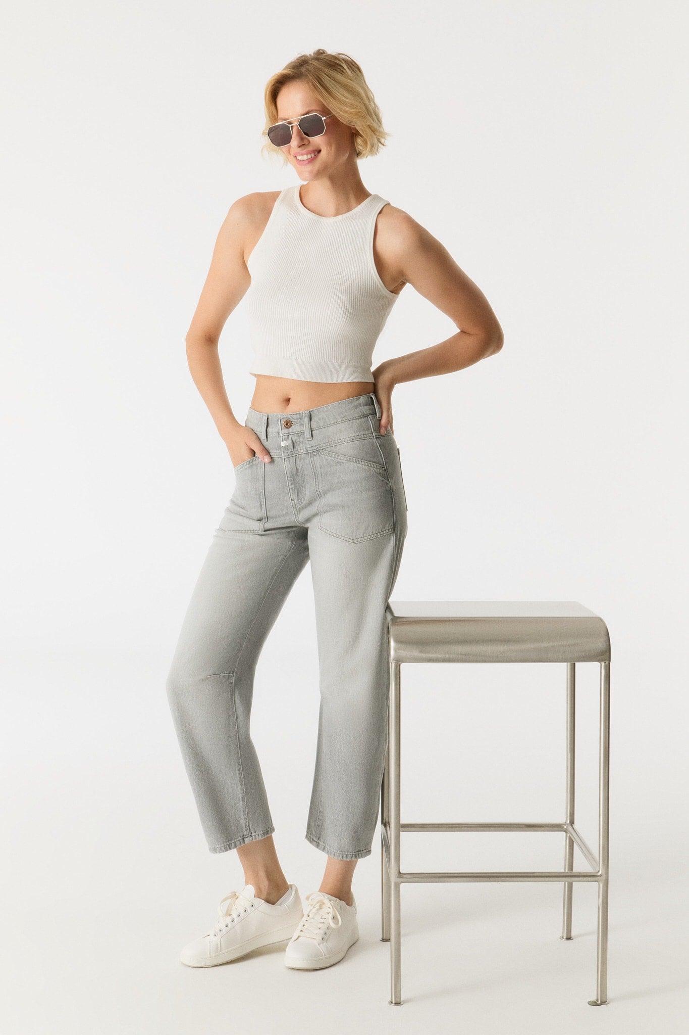 Woman wearing a white sleeveless top and light gray jeans standing next to a stool on a white background